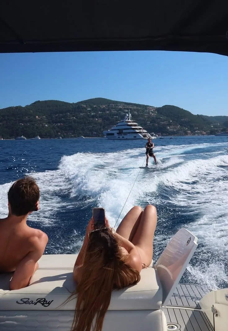 People on a boat watching a person wakeboarding in the water with a large yacht and hills in the background.