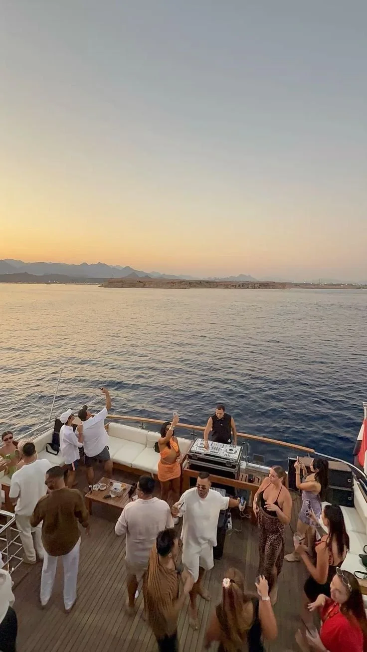 People dancing and socializing on a yacht deck at sunset with a DJ playing music.