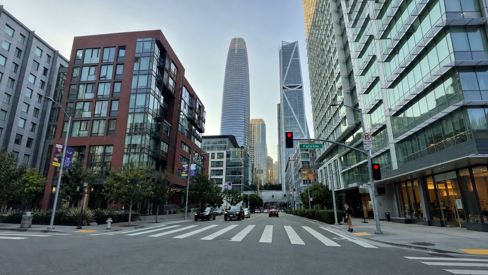 Urban street scene in downtown San Francisco, with modern high-rise buildings, cars, and pedestrians, including a woman crossing the street at a crosswalk. The tallest buildings feature glass facades, and there are traffic signals and street signs, such as one that reads 'Folsom'.