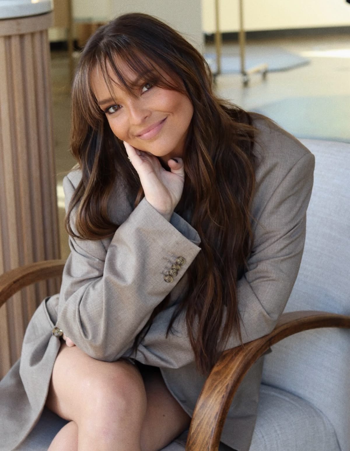 A woman with long brown hair and light skin smiling, resting her face on her hand, sitting on a sofa, wearing a beige blazer.