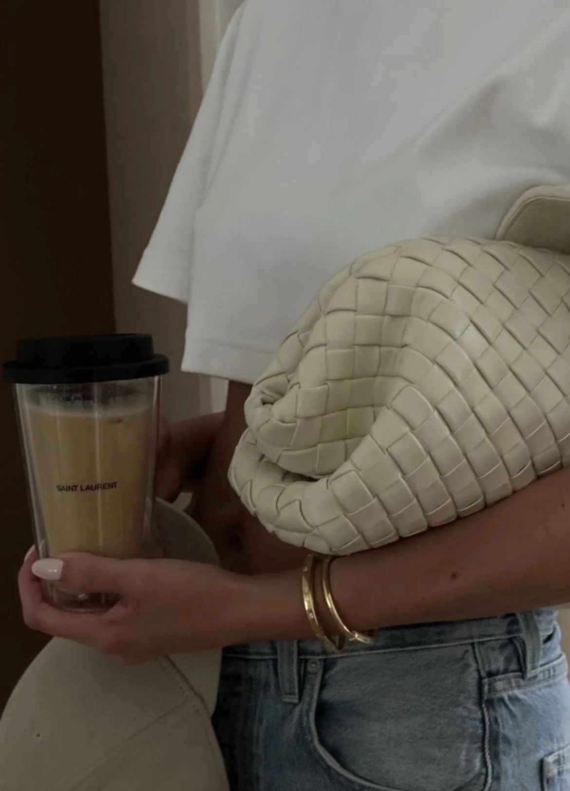 A woman holding a Saint Laurent branded coffee tumbler with a black lid, wearing a white top and jeans, standing next to a beige padded chair.