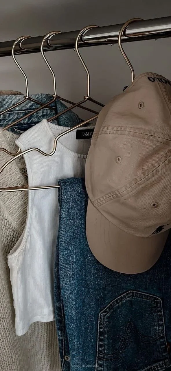Clothing and a beige baseball cap hanging on a closet rod, including white tank top, denim jeans, and a beige long-sleeve shirt.