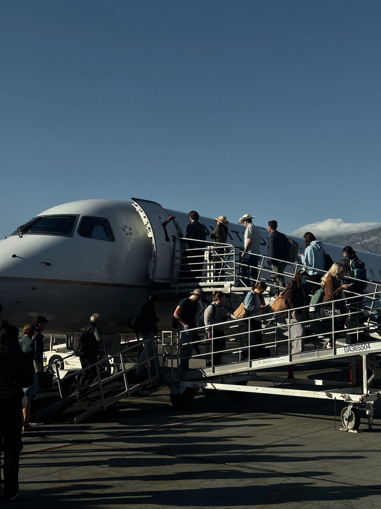 Passengers boarding an airplane via airstairs on the tarmac at an airport, with a mountainous landscape and blue sky in the background.