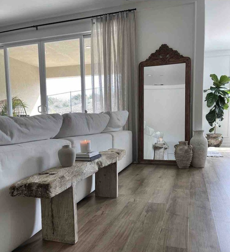 Living room with large white couch, rustic wooden bench, decorative vases, a mirror with ornate wooden frame, and potted plants near sliding glass doors covered with sheer curtains.
