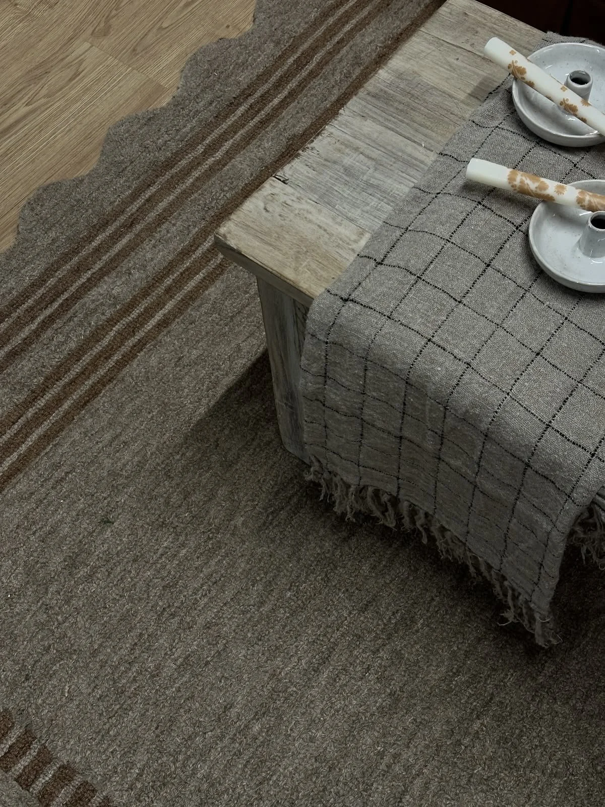 A wooden table with a linen tablecloth, two white candlestick holders with decorative candles, and a brown and beige striped and checkered area rug on a hardwood floor.