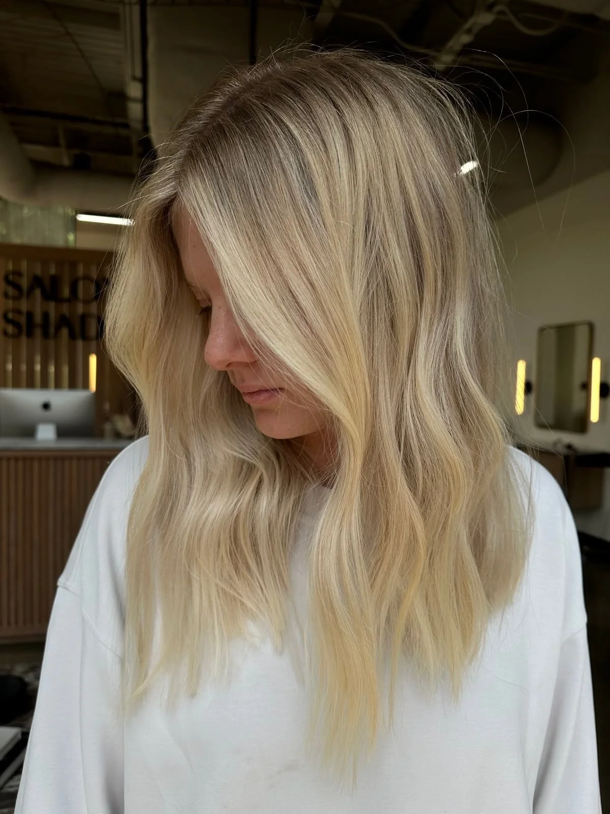 A woman with long, wavy blonde hair looking down, inside a hair salon.