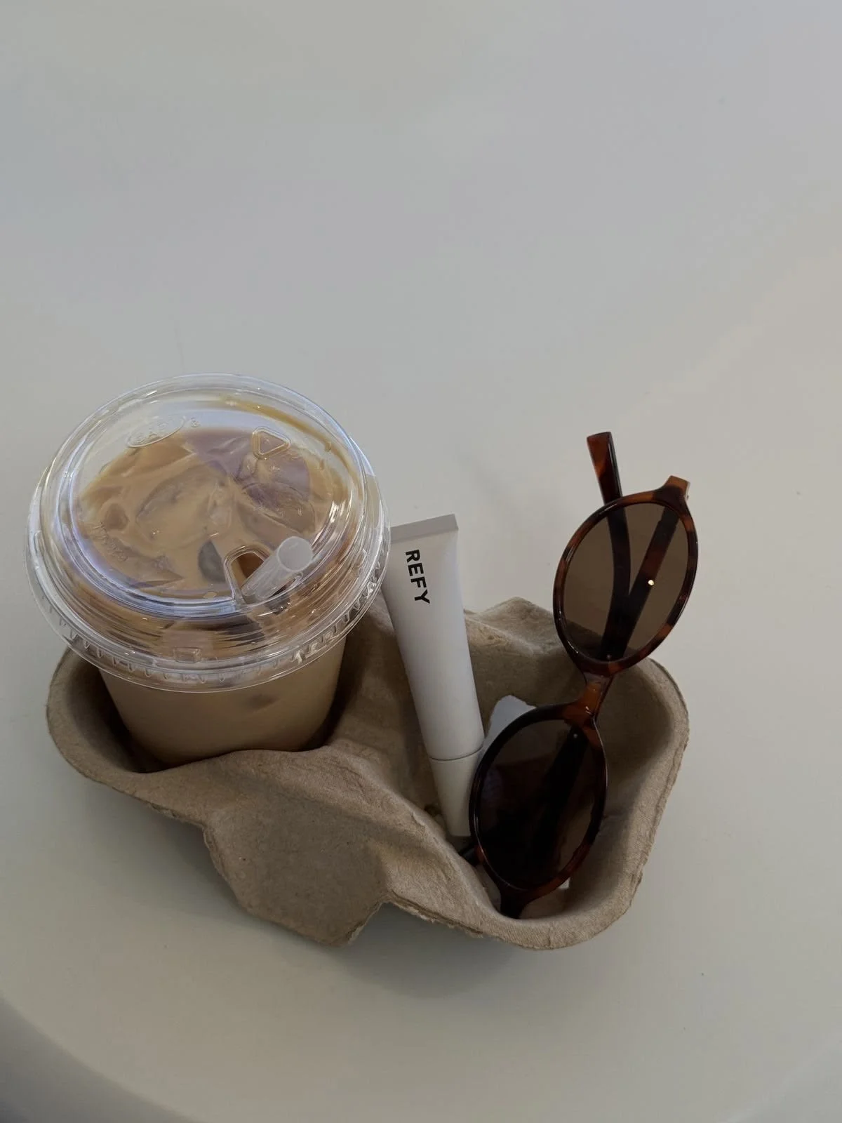 A takeaway iced coffee in a plastic cup with a clear lid and straw, a pair of tortoiseshell sunglasses, a white lipstick tube labeled 'REFY', all placed in a beige fabric holder on a white surface.