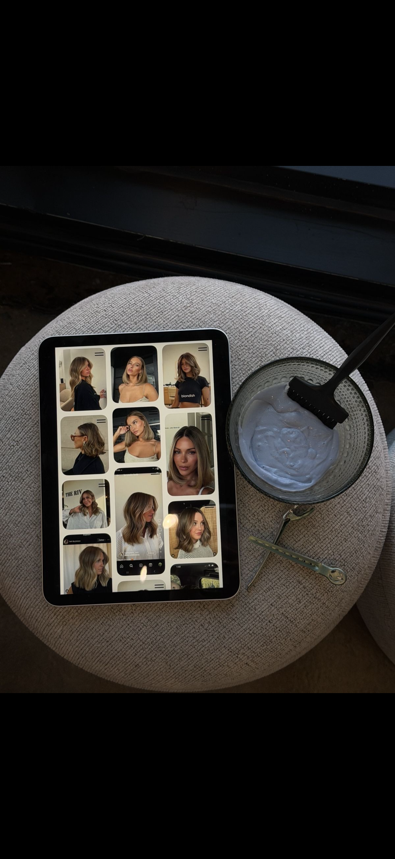 A tablet displaying photos of a woman with blonde hair styled in various ways. Next to the tablet is a glass of white yogurt or smoothie with a spoon and a black hairbrush. The items are on a round, textured beige cushion.