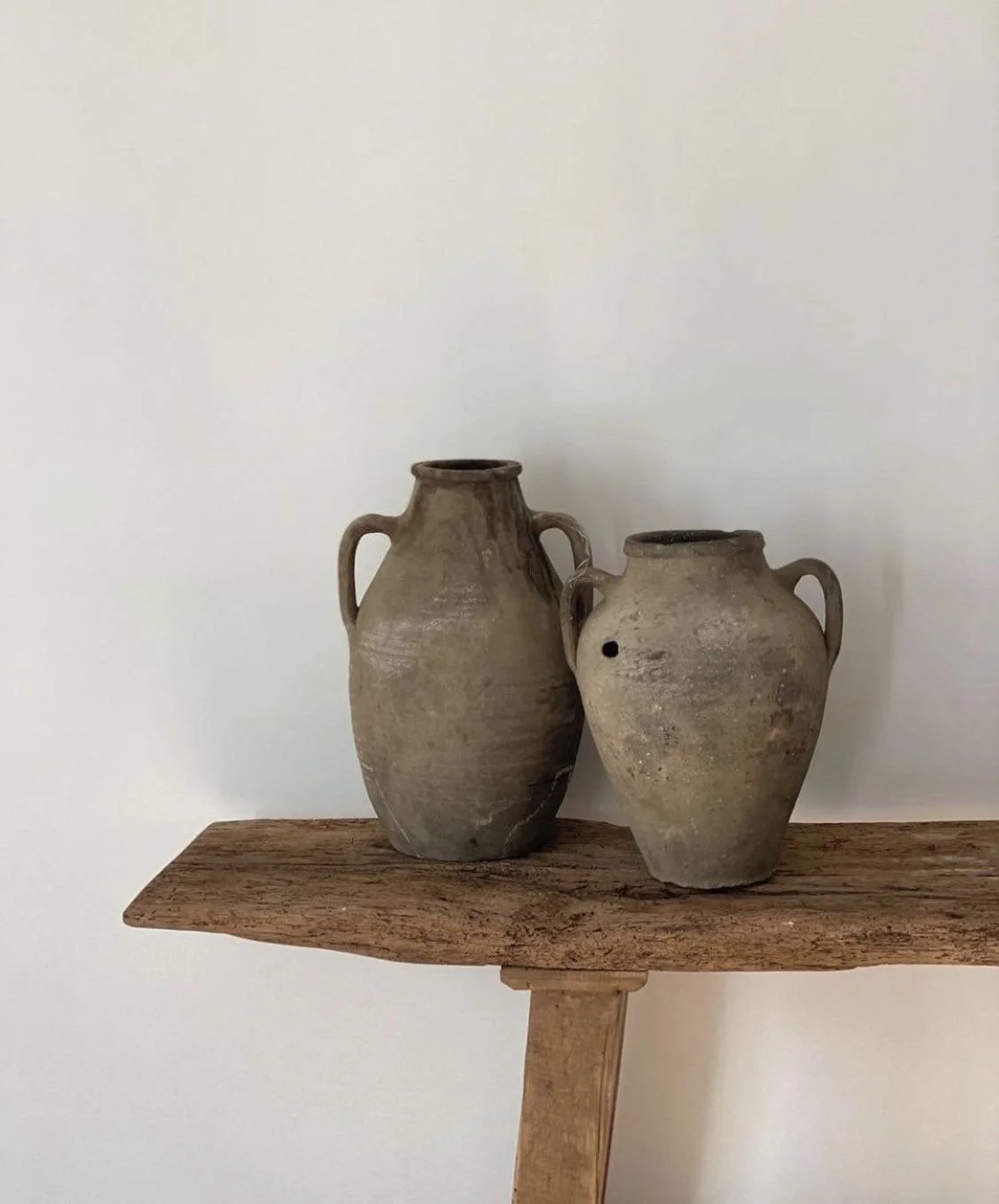 Two ancient clay pots with handles on a weathered wooden shelf against a plain white wall.