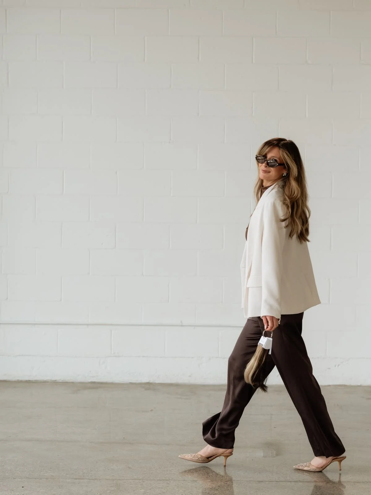 A woman with long wavy hair wearing dark sunglasses, a white blazer, dark trousers, and nude high heels, walking indoors against a plain white brick wall.