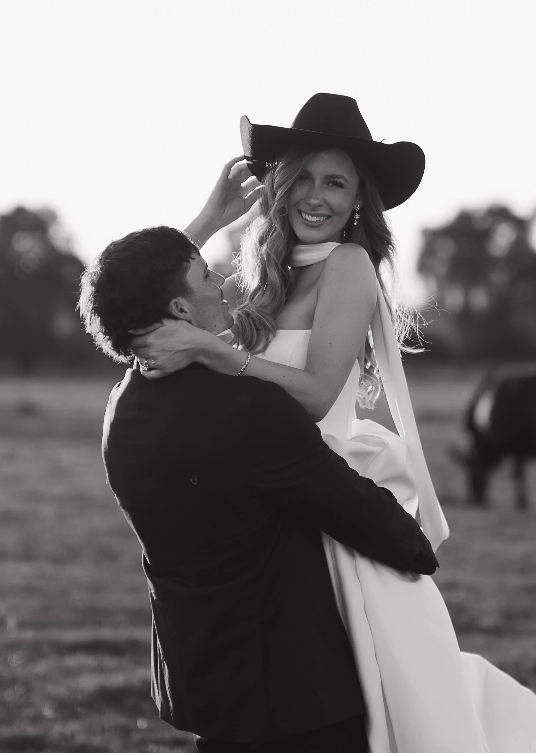 A black-and-white photograph of a couple at sunset, with the man lifting the woman in his arms. The woman is smiling and wearing a large hat, a sleeveless dress with a sash around her neck, and elegant earrings. The man is looking up at her with his arms around her, in a dark suit. The background features trees and an open field.
