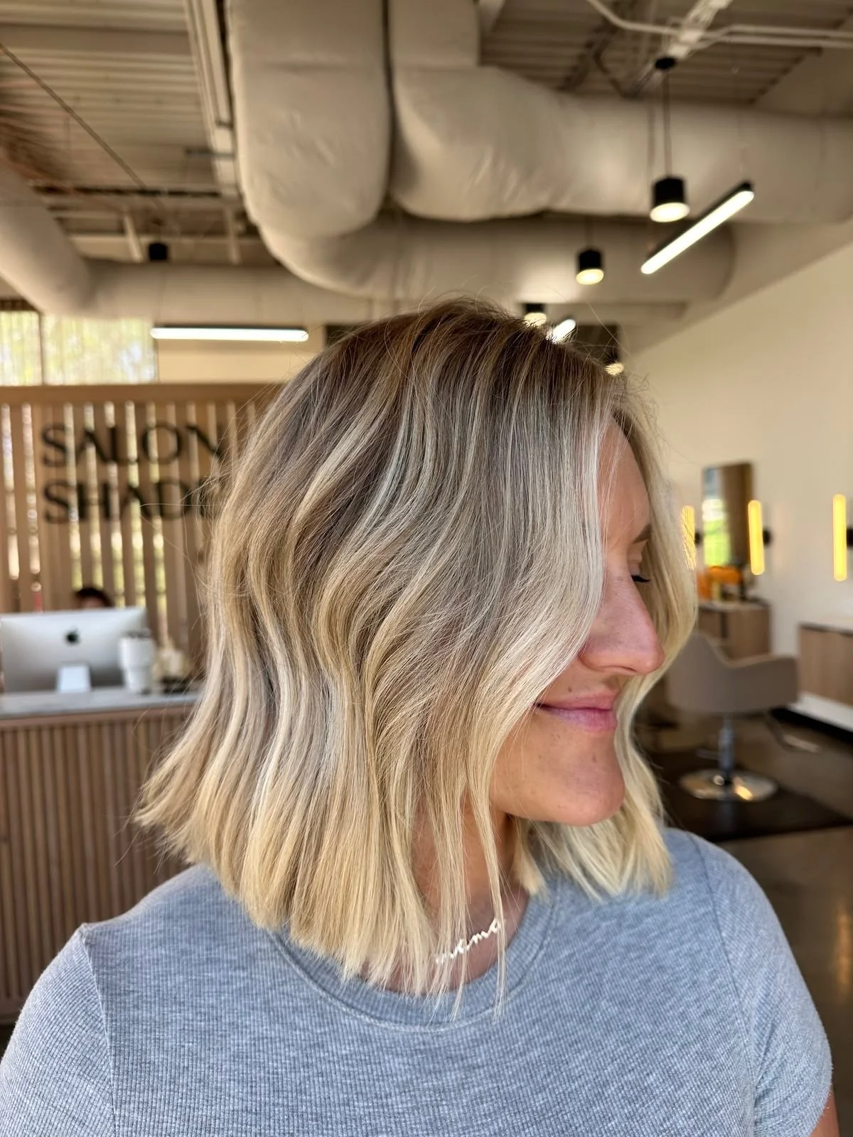 A woman with shoulder-length blonde hair with dark roots, styled in loose waves, smiling with her eyes closed, in a salon setting with modern decor.