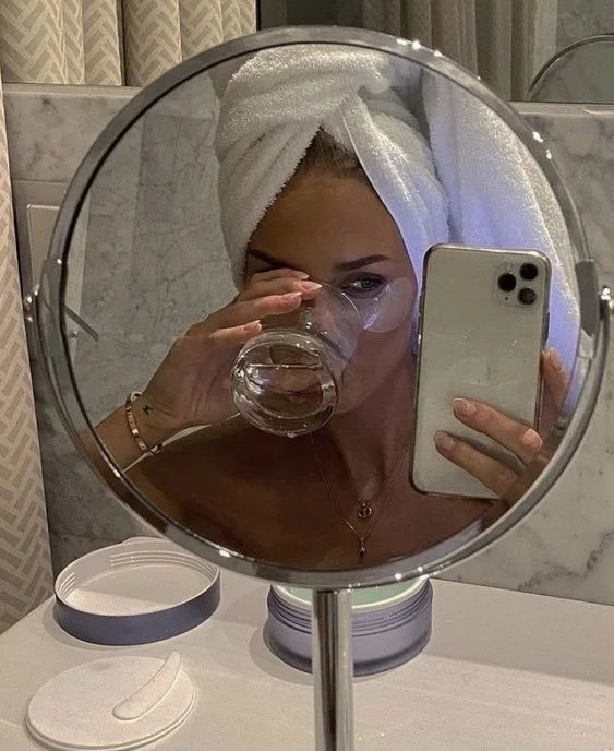 Woman with a towel wrapped around her head taking a selfie in a bathroom mirror, drinking water from a glass, holding a smartphone in her right hand.