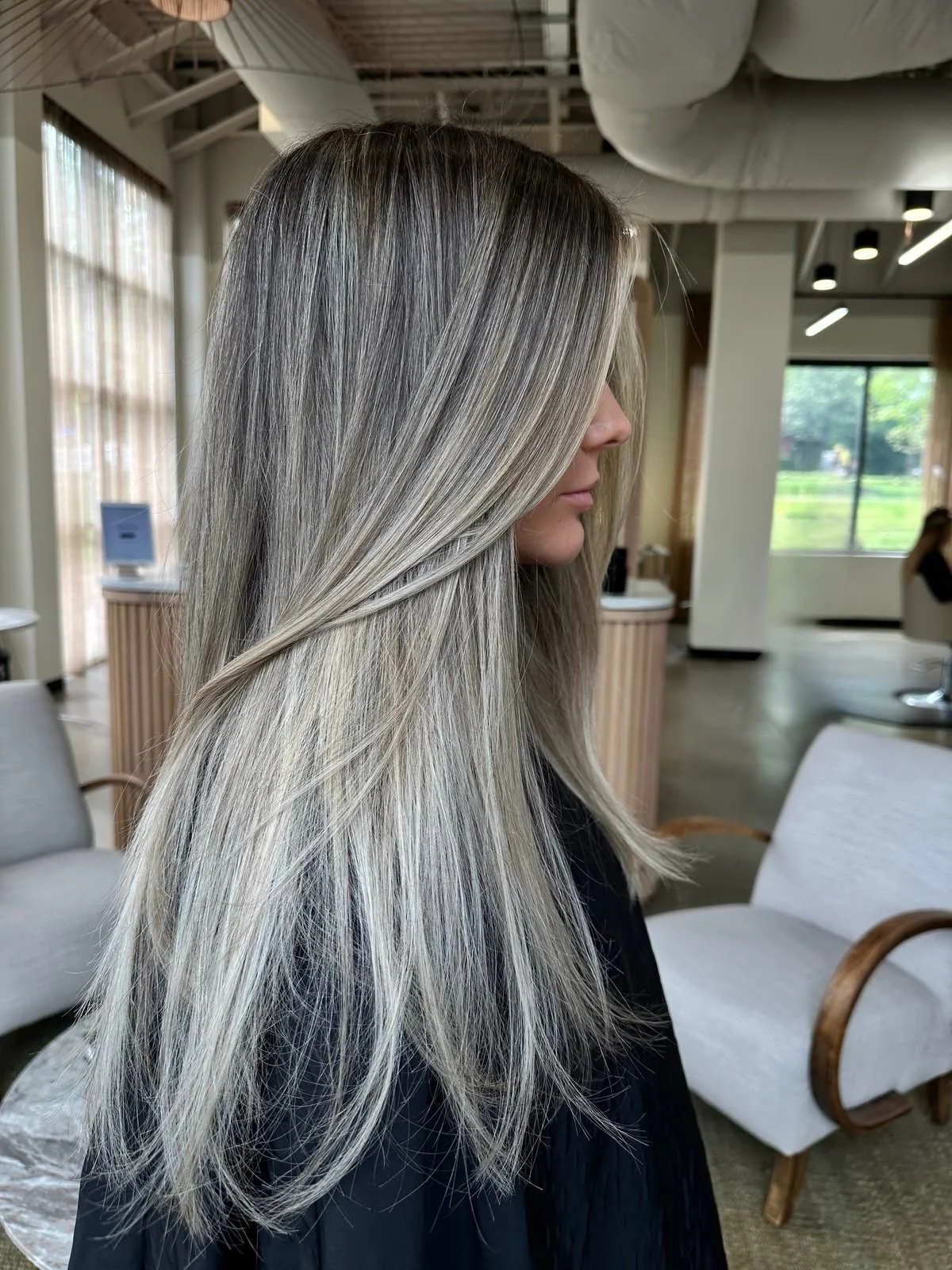 Side profile of a woman with long, blended ash blonde hair in a salon with natural light, chairs, and large windows.