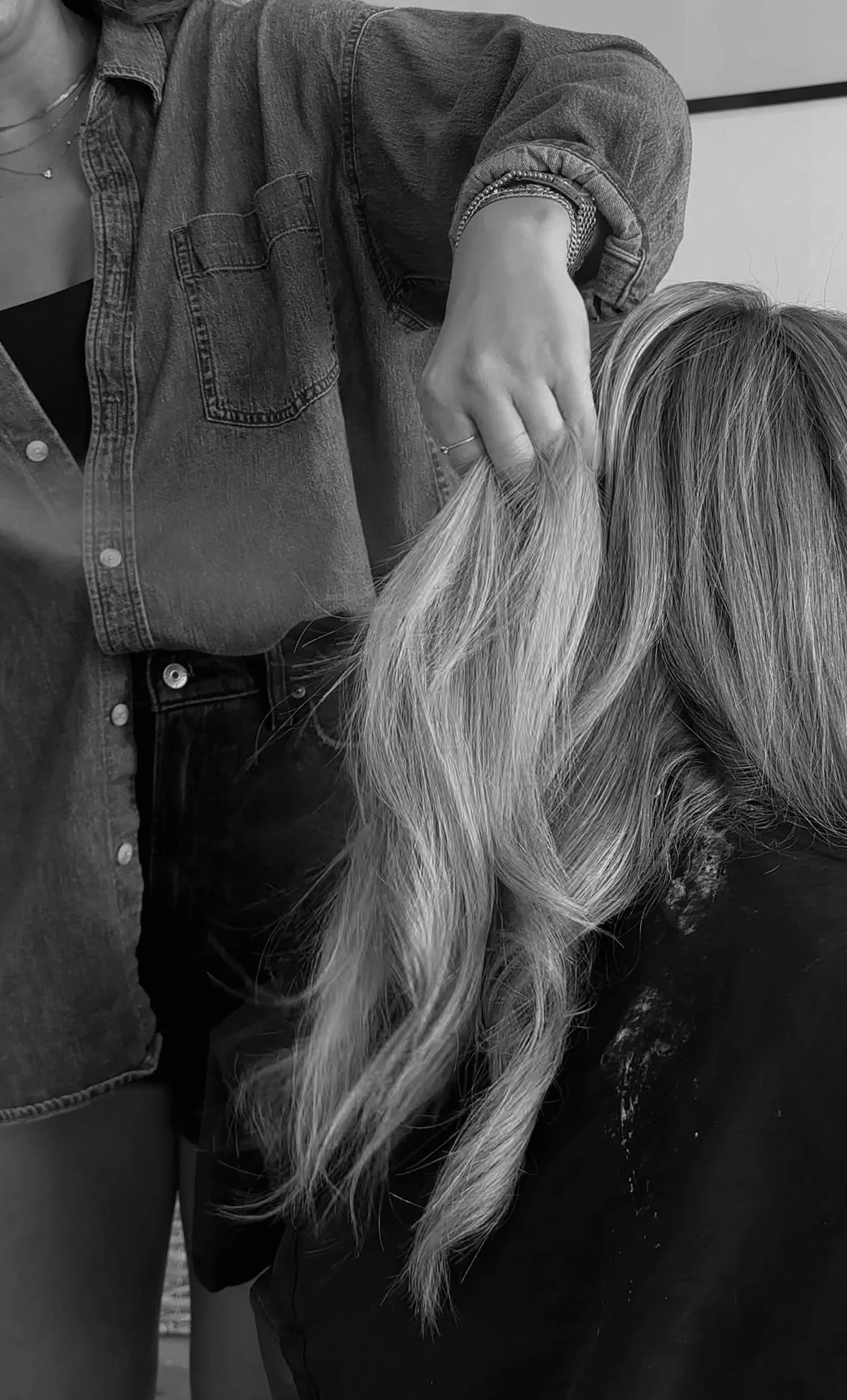 A person with long, wavy blond hair is being touched on the head by another person wearing a denim jacket. The image is in black and white.