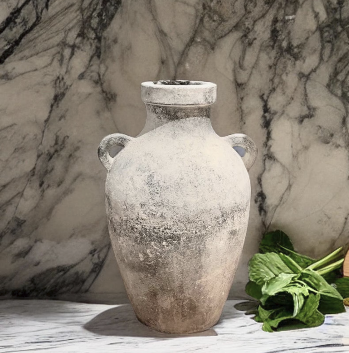 An ancient stone vase with two small handles, weathered and aged, placed on a marble surface, with fresh green mint leaves next to it against a marble wall background.