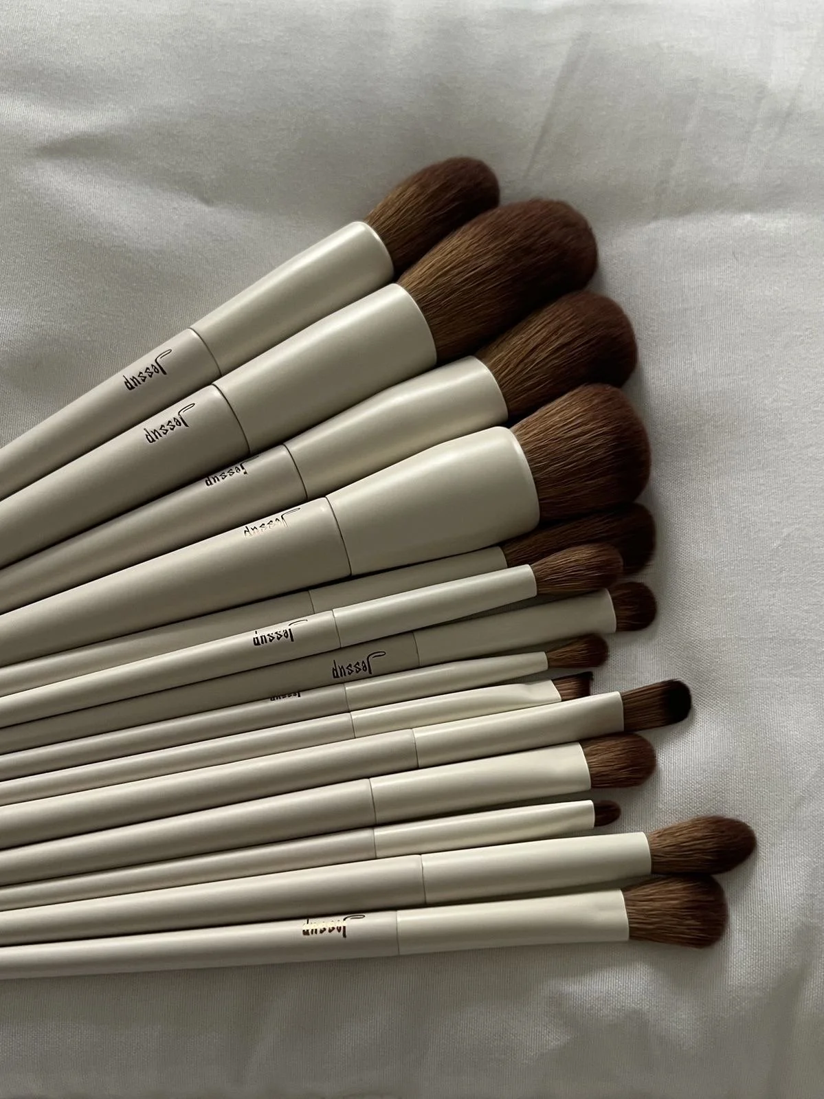 A collection of makeup brushes with light handles and brown bristles, arranged on a white surface.