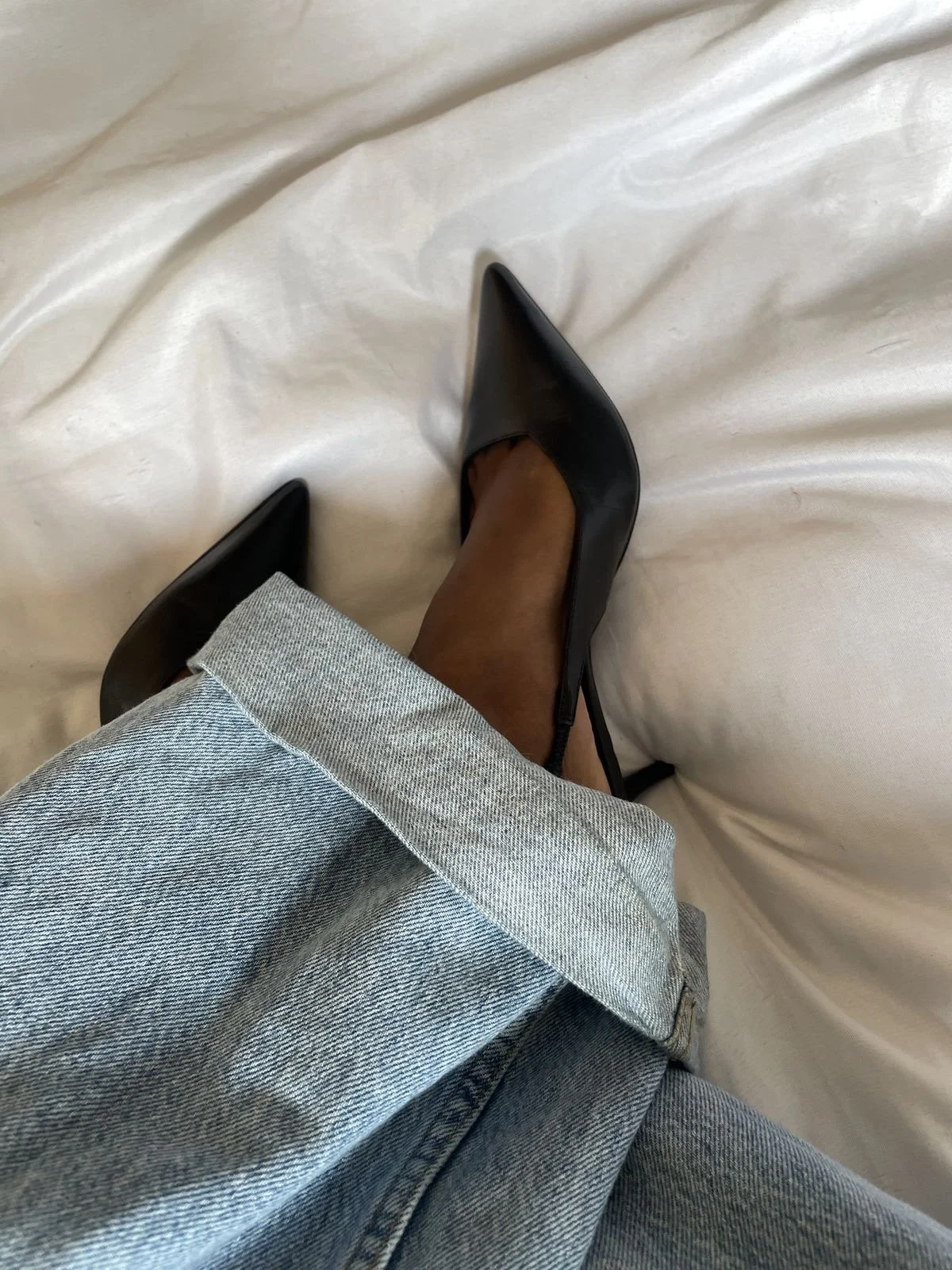 Close-up of a person's legs and feet, wearing gray jeans with rolled cuffs and black pointed high heels, resting on a white sheet.