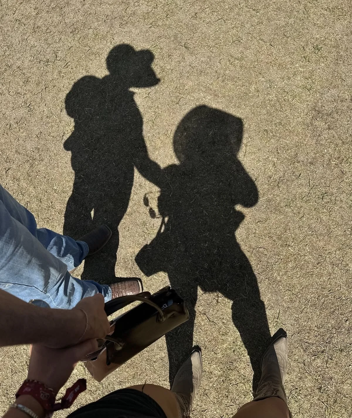 Shadows of two people holding hands and walking, with one person carrying a bag, cast on a sandy ground.
