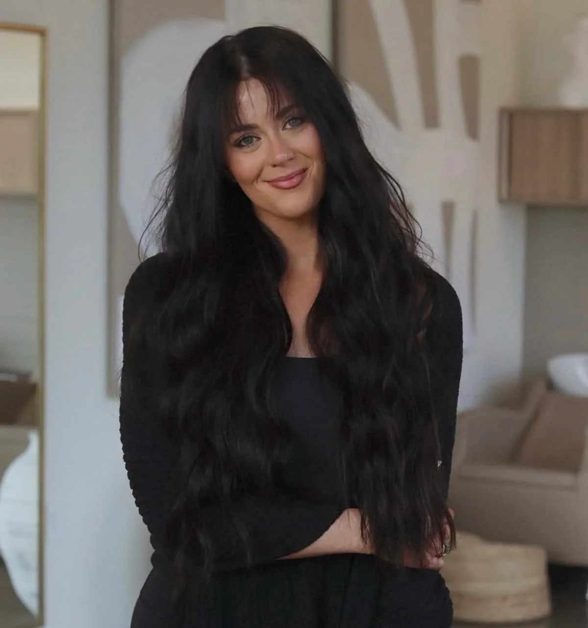 A woman with long black wavy hair, wearing a black top, smiling, standing indoors in front of a neutral wall and modern decor.