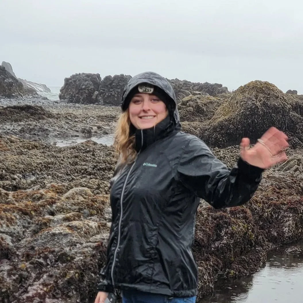 Smiling woman waving at rocky, tide-covered shore in rainy weather, wearing a black waterproof jacket and a helmet.