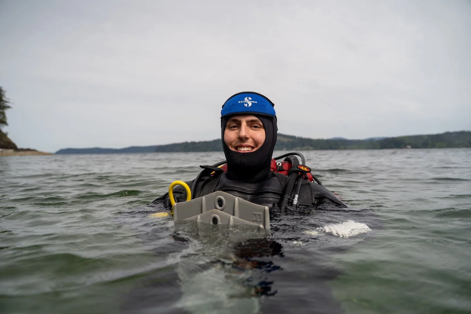 A person in scuba diving gear smiling while floating in the water with a distant shoreline and cloudy sky in the background.