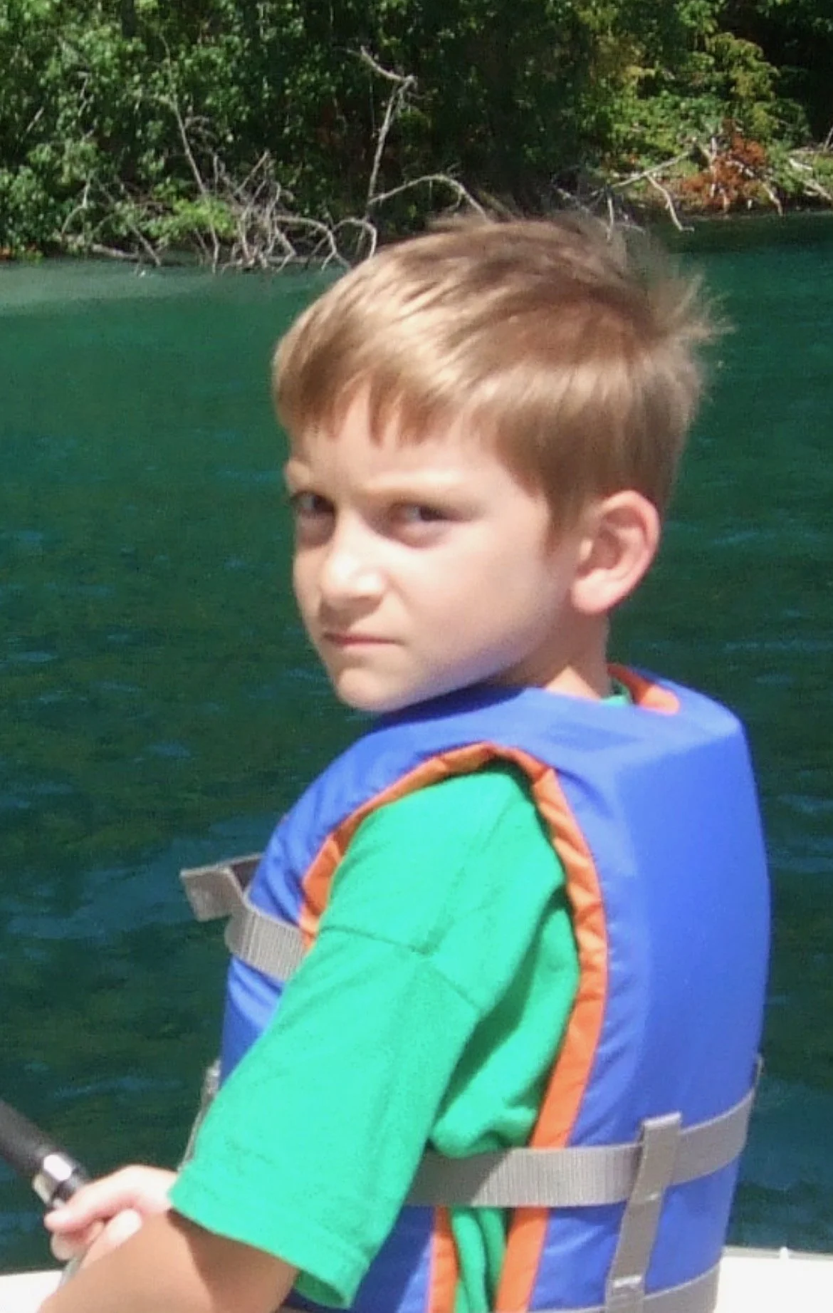 A young boy with short light brown hair and a serious expression, wearing a green shirt and a blue life vest, is on a boat near a body of water with trees and bushes in the background.