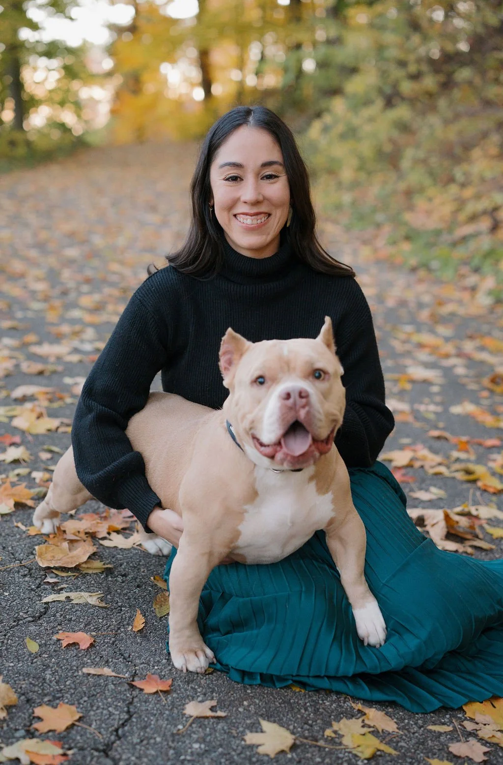 A woman in a black sweater and teal skirt sitting outdoors on a path surrounded by fallen autumn leaves, smiling with a tan and white dog on her lap.