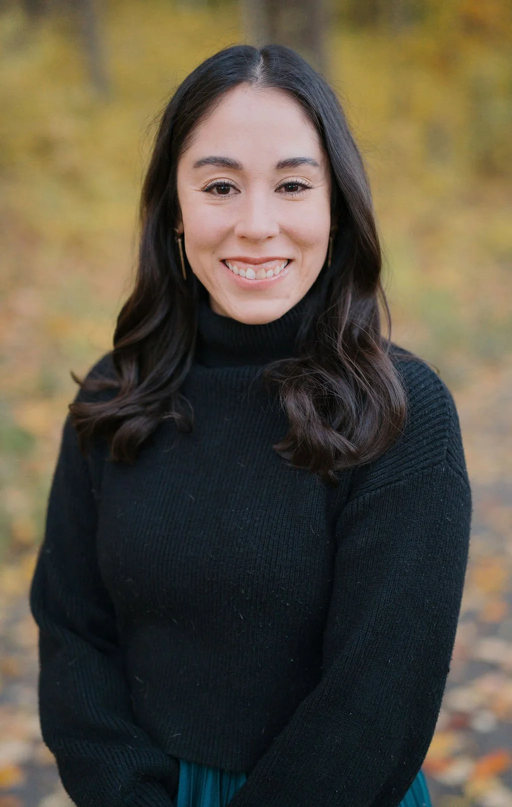 A young woman with long dark hair smiling outdoors in a black turtleneck sweater with fall foliage in the background.