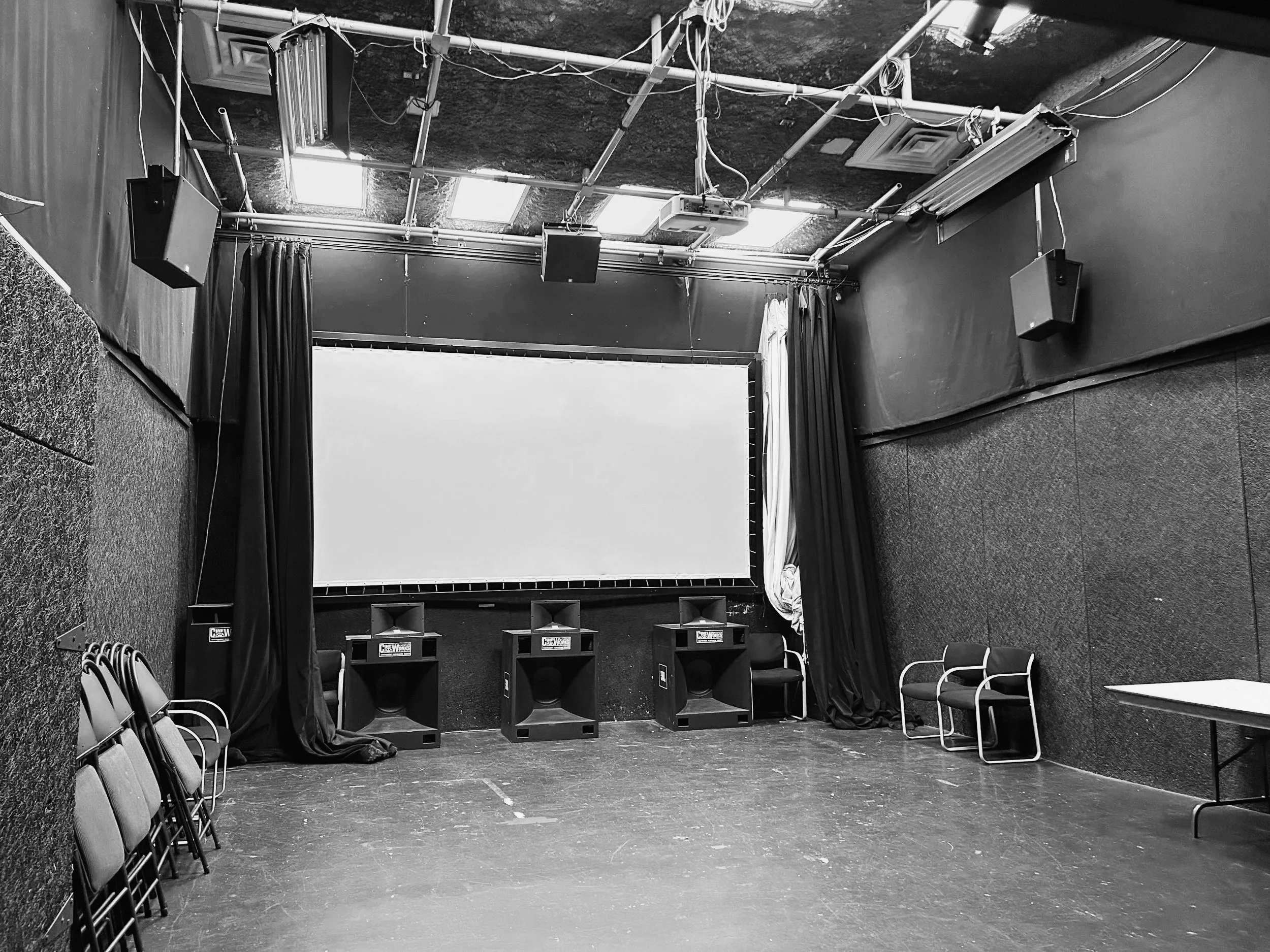 Empty film studio with chairs arranged on the side, black curtains, speakers, and lighting equipment in a small theater or conference room.