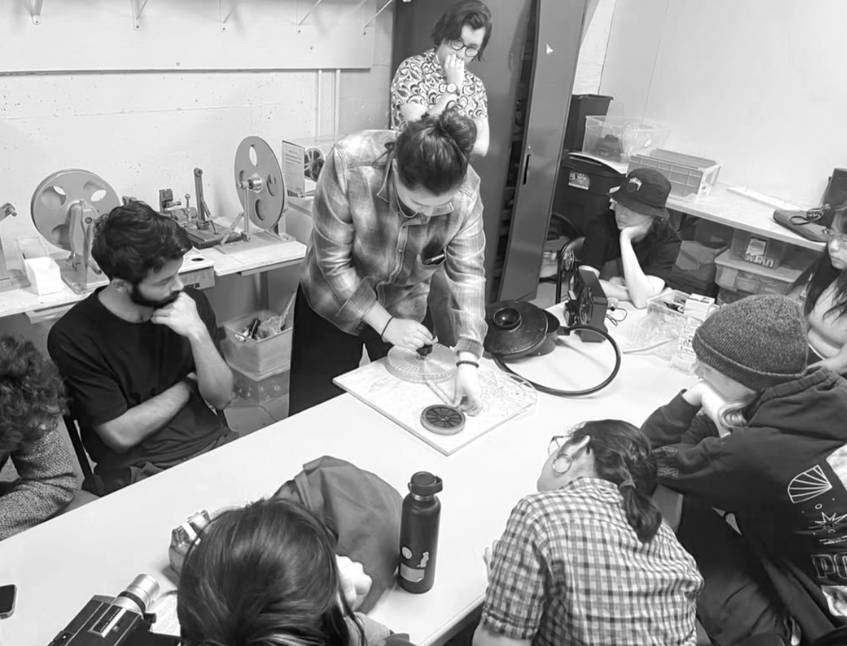 A group of people gathered around a table in a workshop, with a woman demonstrating an experiment or activity at the center. Others are watching attentively, some with thoughtful or curious expressions.