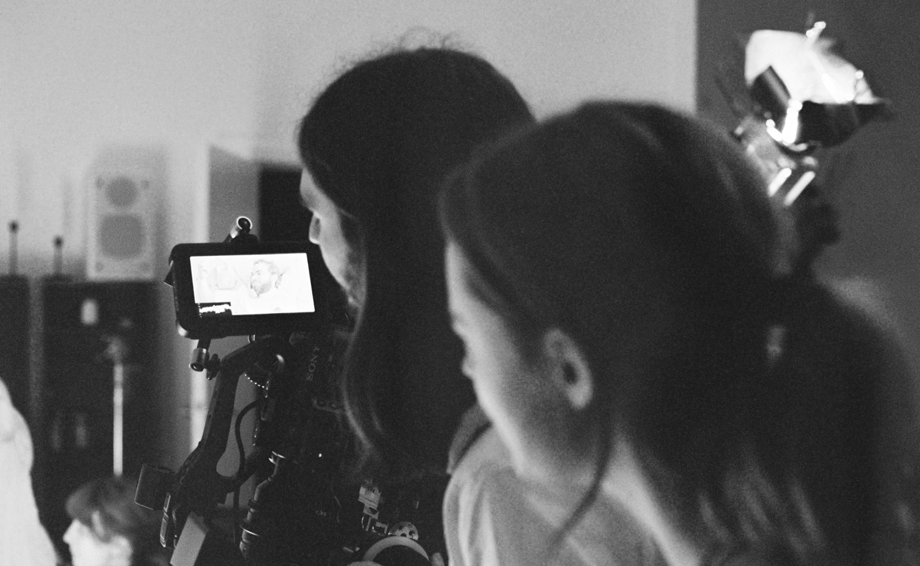 Black and white photograph of a film set with a camera on tripod, two women, and a woman wearing a headscarf, all facing away from the camera, with studio lights in the background.