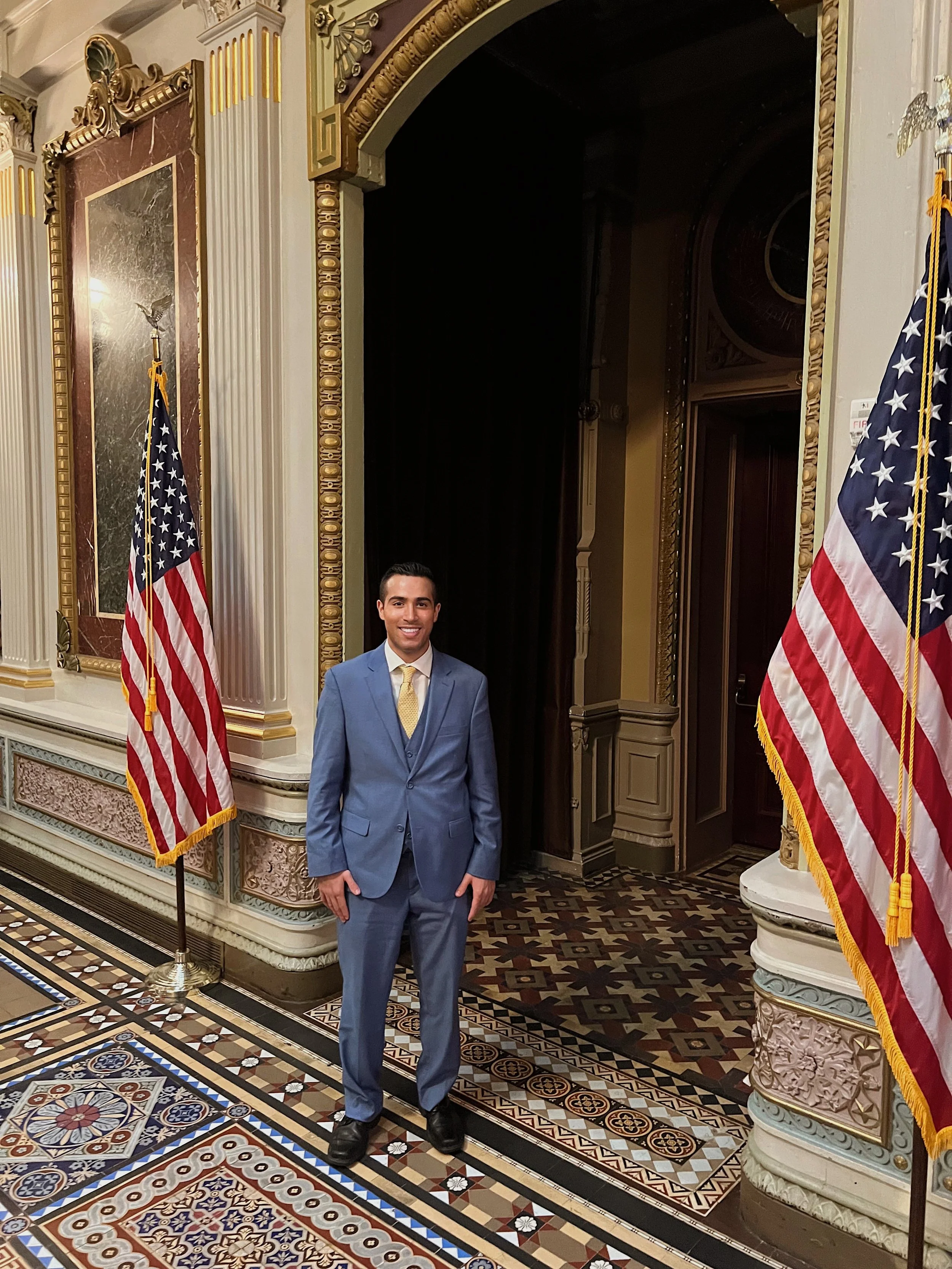 A man wearing a light blue suit and yellow tie standing indoors in front of an ornate wall with gold and marble accents. There are two American flags on either side of him, and intricate patterned tile flooring beneath him.