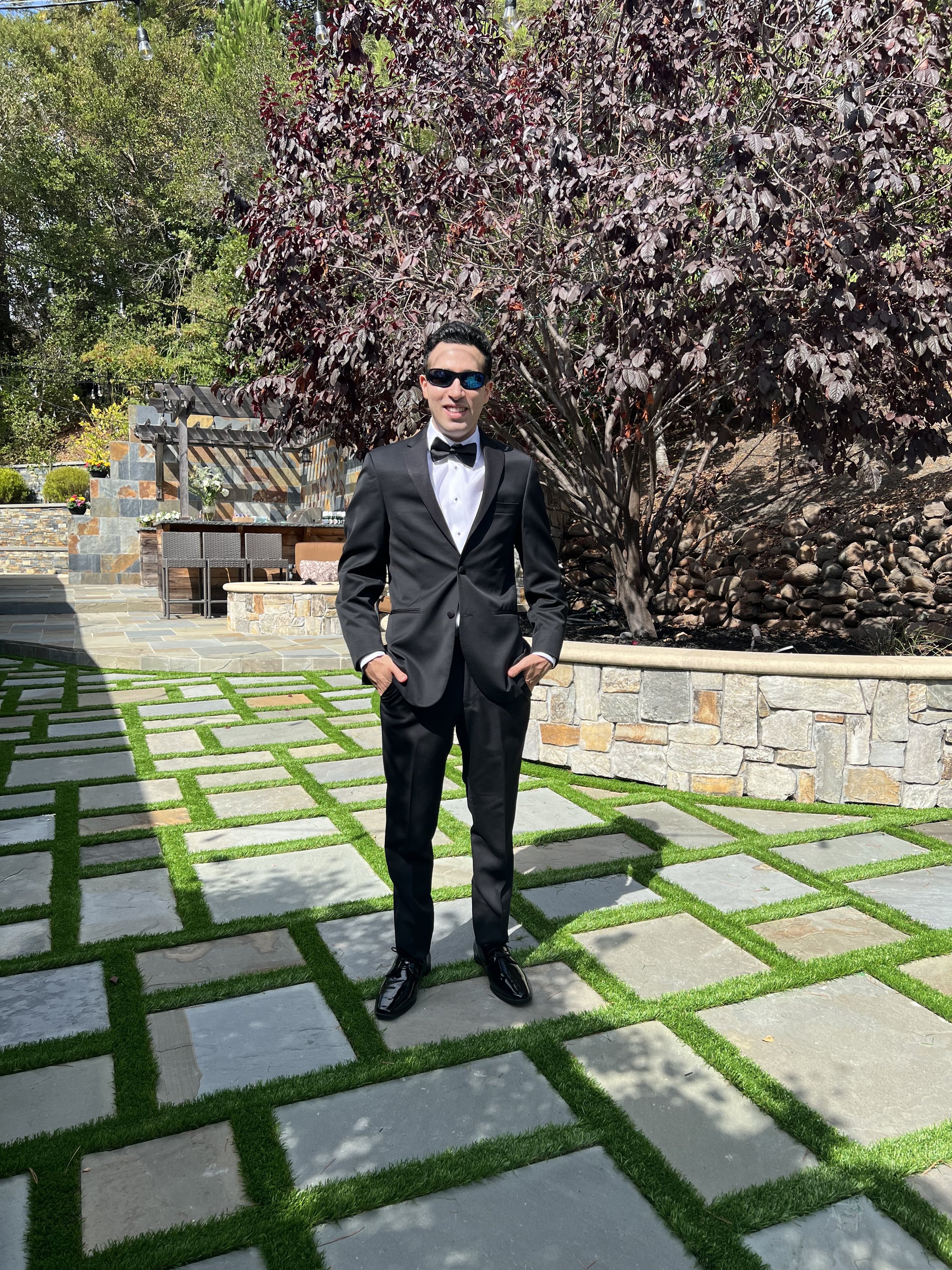 A man in a black tuxedo and sunglasses standing outdoors on a patterned stone and grass walkway, with a large dark red-leafed tree and outdoor furniture in the background.