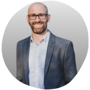 A smiling man with glasses, a beard, wearing a suit jacket and a collared shirt, standing against a plain background.