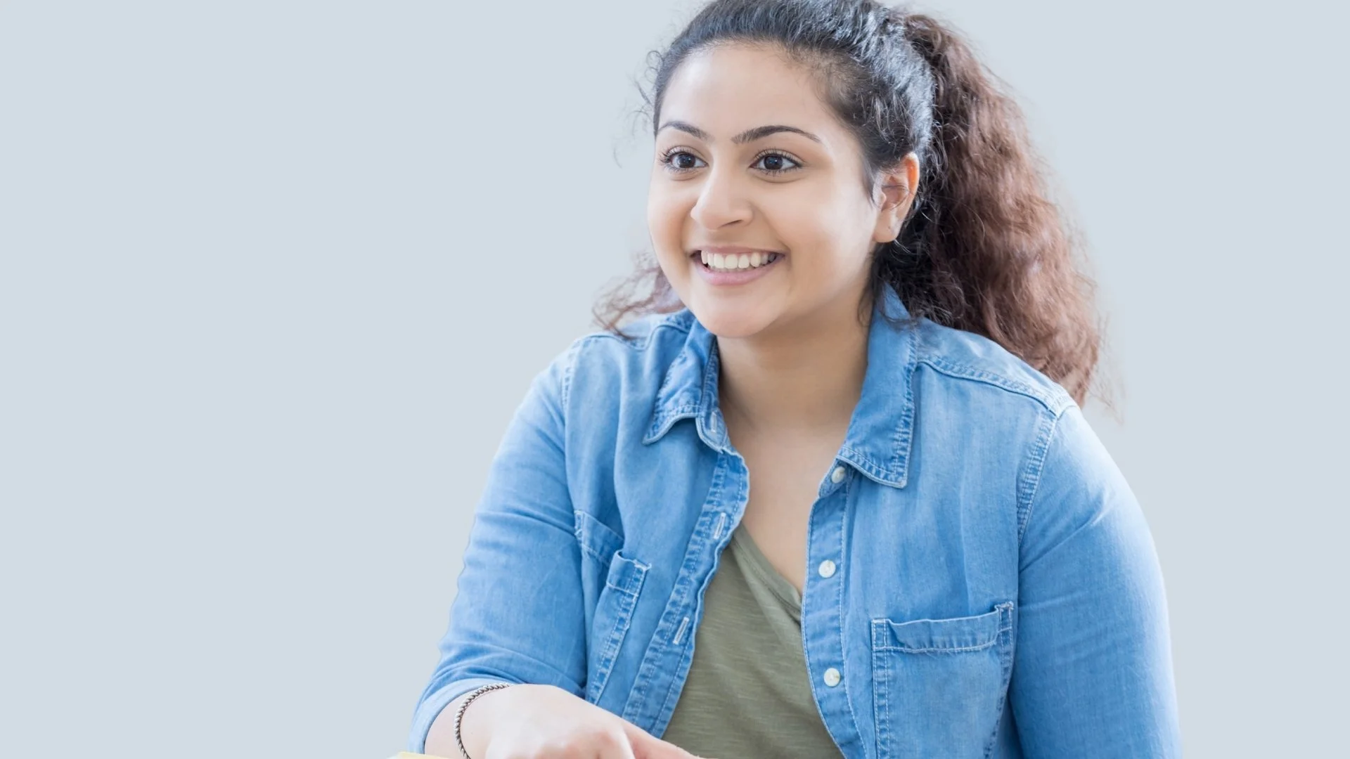 A young woman with curly hair tied back, wearing a denim shirt, smiling and looking to the side.