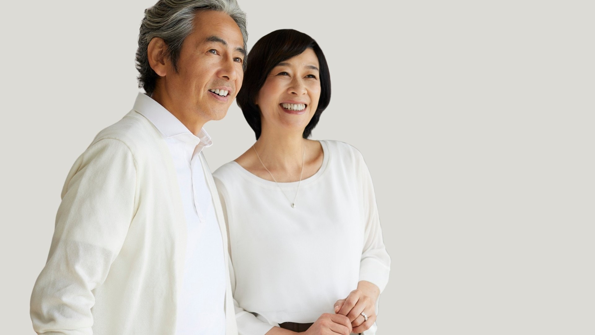 A middle-aged Asian man and woman smiling and standing together against a plain light-colored background.