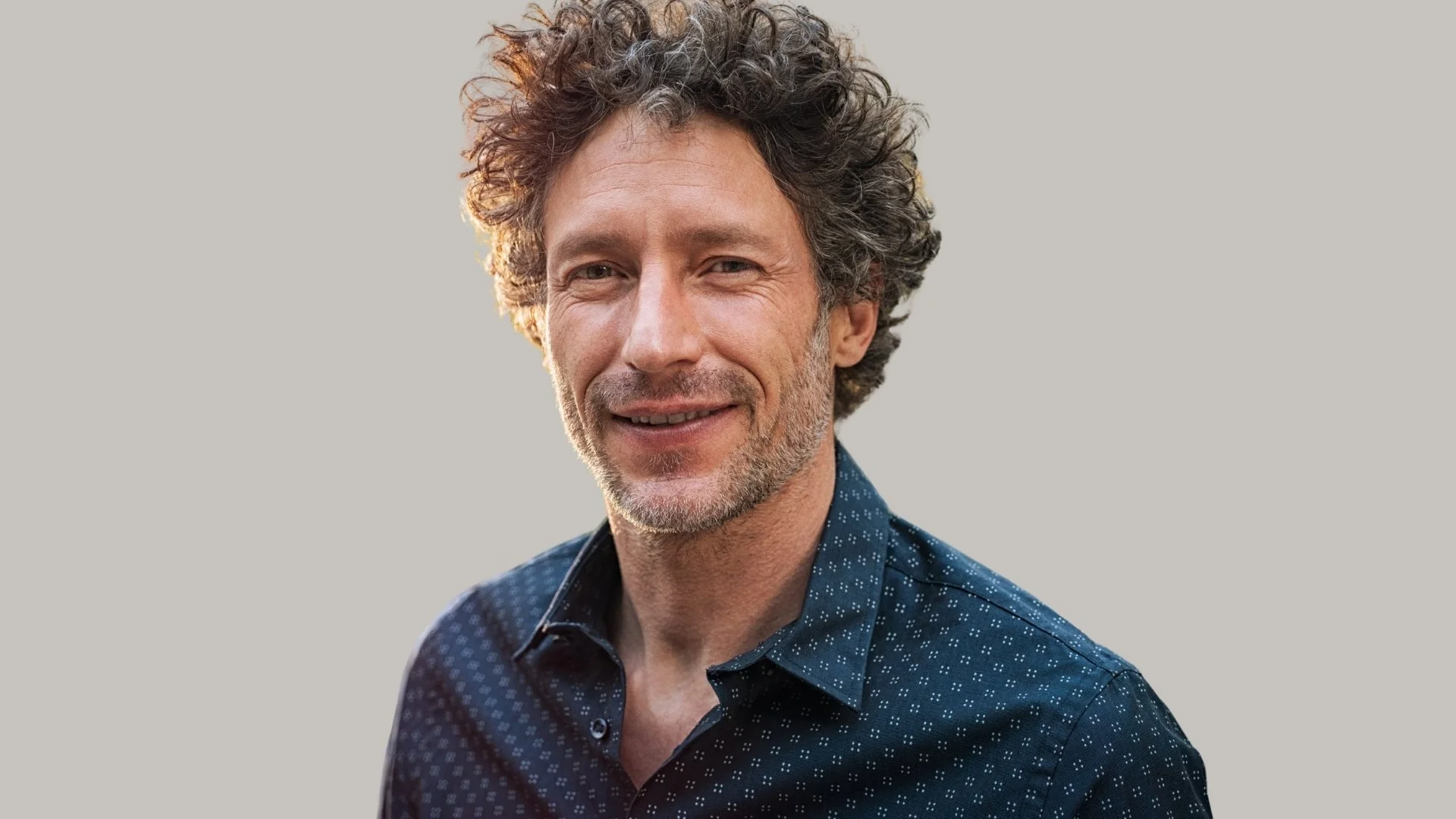 Close-up of a smiling middle-aged man with curly hair and a beard, wearing a dark blue patterned shirt, against a plain light background.