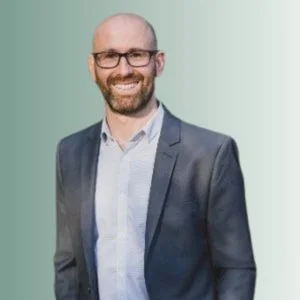 A smiling man with glasses, a beard, and a bald head, wearing a gray suit jacket over a light blue shirt, standing against a light green background.