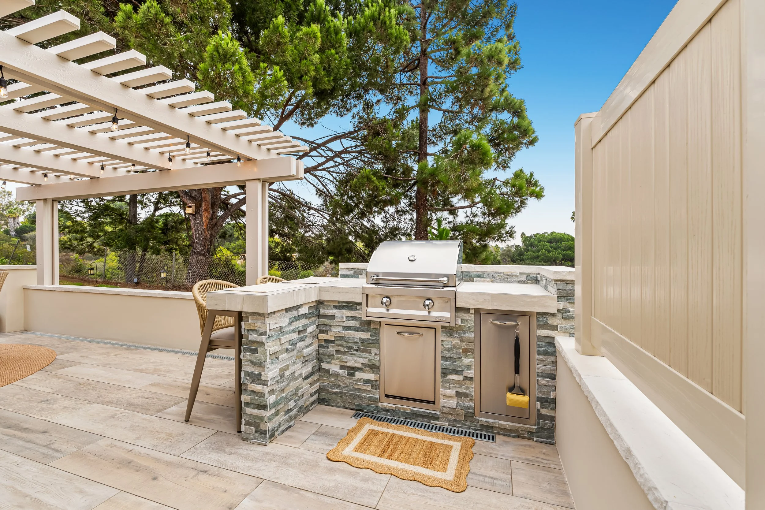 Outdoor kitchen area with a built-in gas grill, cabinetry, and a countertop made of stone. There is a wooden trellis overhead with string lights, and green trees in the background. The area has beige flooring and a small beige and orange rug in front of the grill.