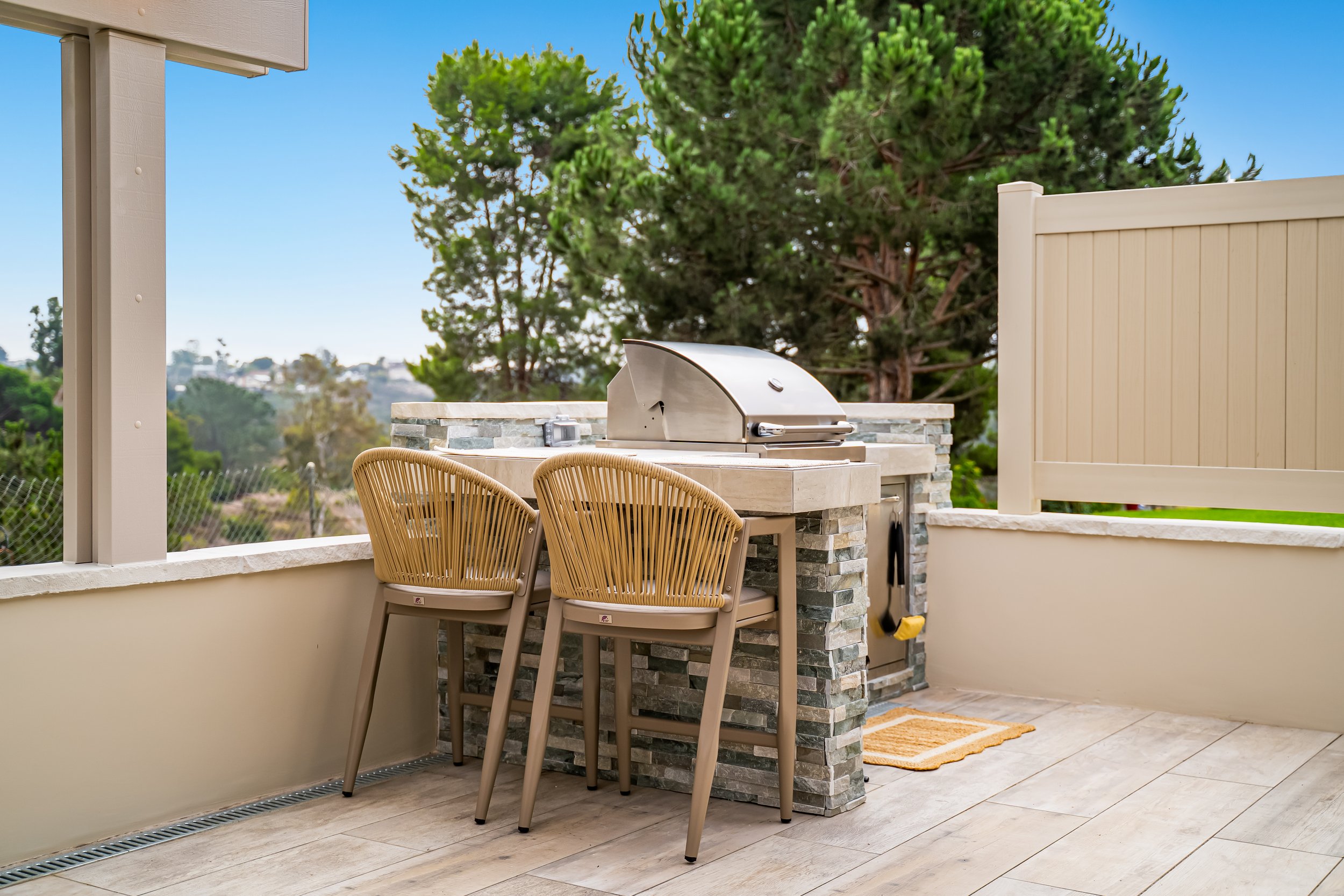Outdoor patio area with a built-in stainless steel propane grill, four beige woven chairs, a stone counter, a beige privacy fence, and lush green trees with a clear blue sky in the background.
