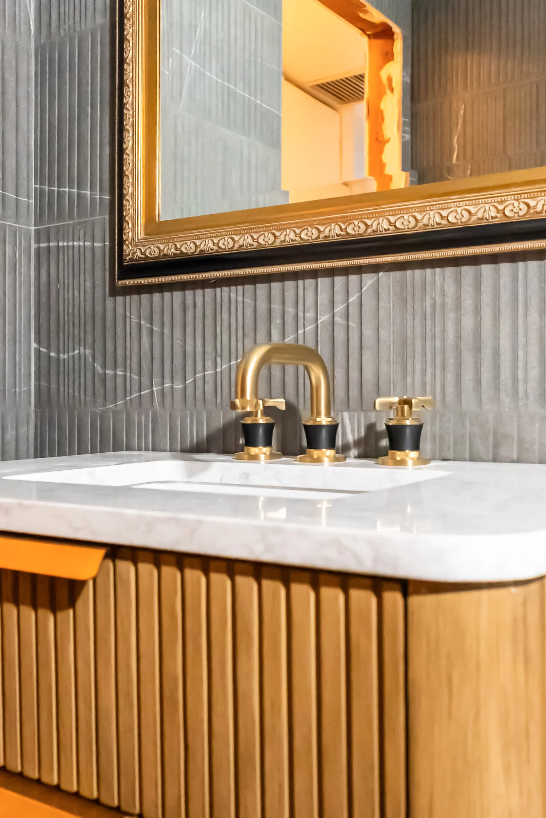 A bathroom sink with a black and gold faucet, a marble countertop, a large ornate gold-framed mirror, dark textured wall tiles, and a wooden cabinet underneath.