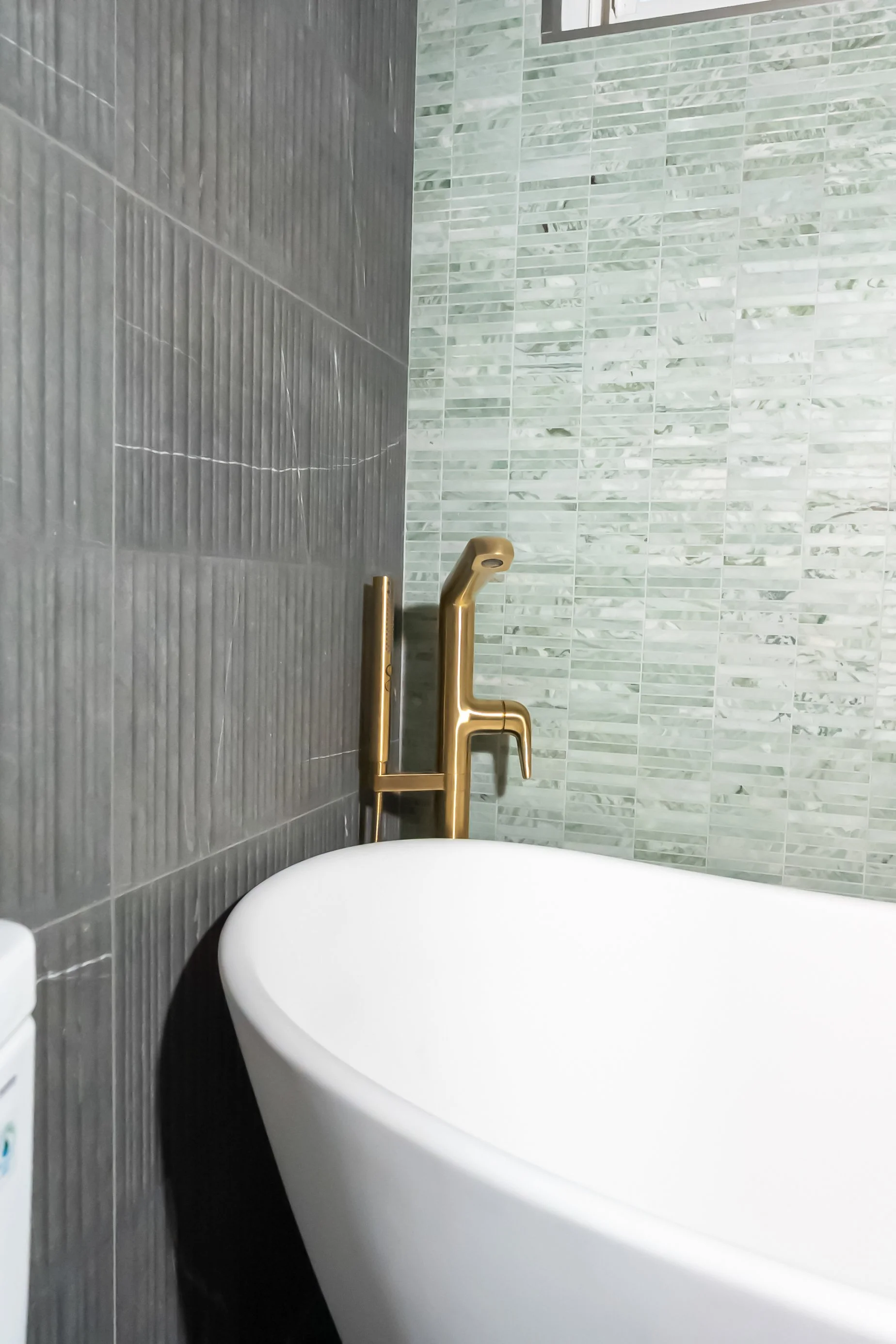 Close-up of a modern bathroom with a white freestanding bathtub, a gold faucet, and tile walls in shades of gray and green.