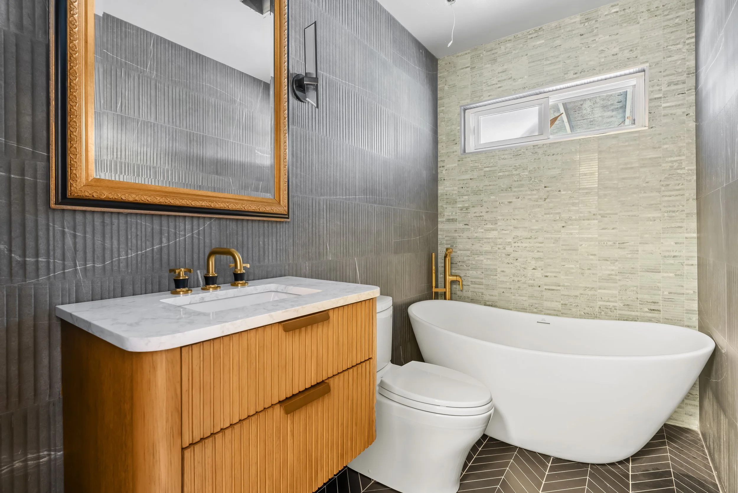 Modern bathroom with gray tiled wall, a freestanding white bathtub, a wooden vanity with a marble countertop, and a large mirror with an ornate gold frame. There's a small horizontal window on the tiled wall.