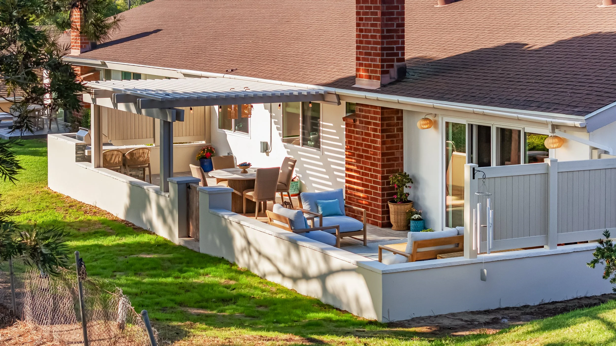 A house with a patio and outdoor furniture, including chairs and a table, surrounded by a white fence. The house has a red brick chimney, beige paneling, and large windows, with decorative lanterns hanging outside. The yard is grassy with some trees and a chain-link fence in the foreground.