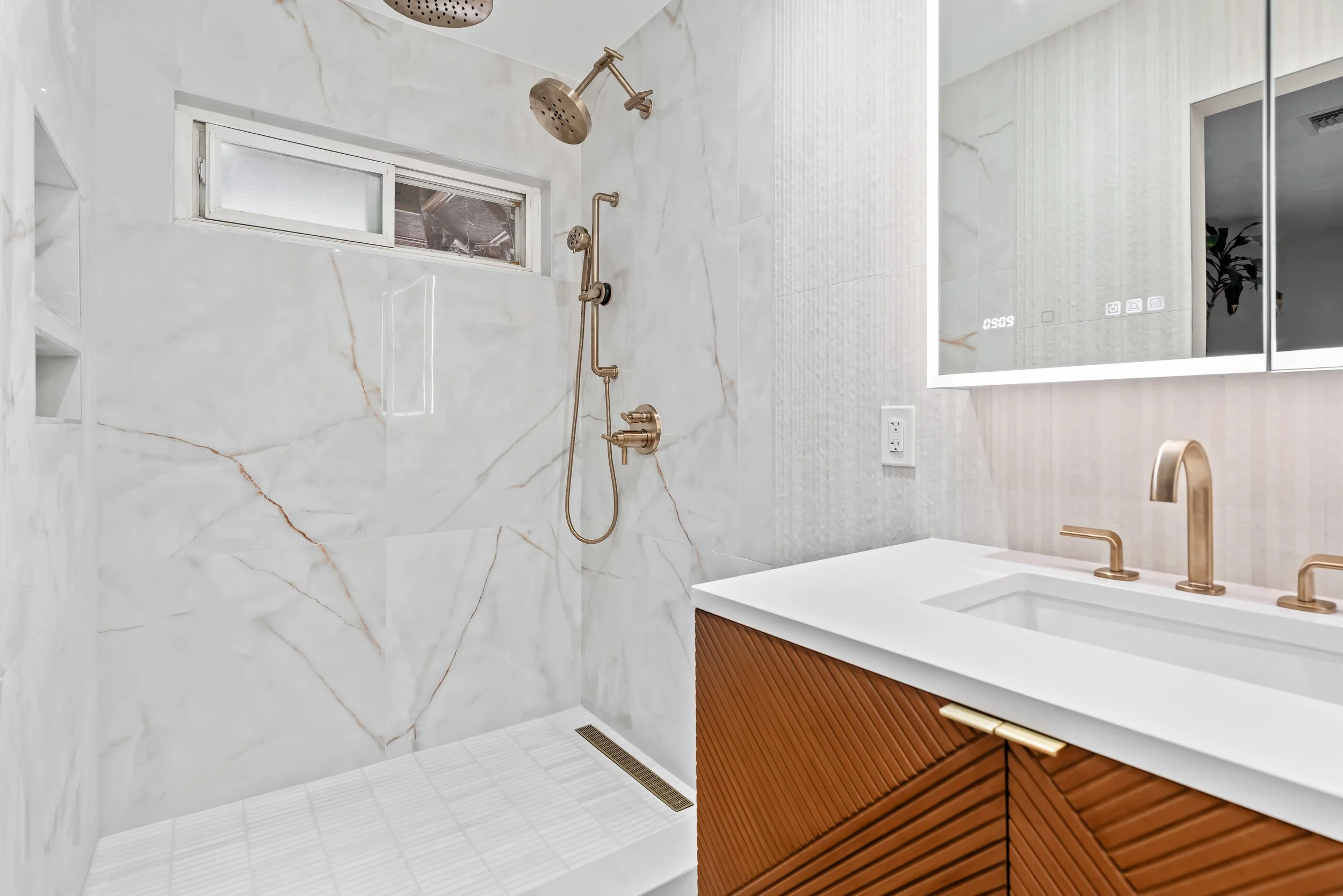 Modern bathroom with a marble-tiled shower area, a small window, gold fixtures including a showerhead and faucet, and a wooden vanity with a white countertop and a gold faucet, reflected in a large backlit mirror.