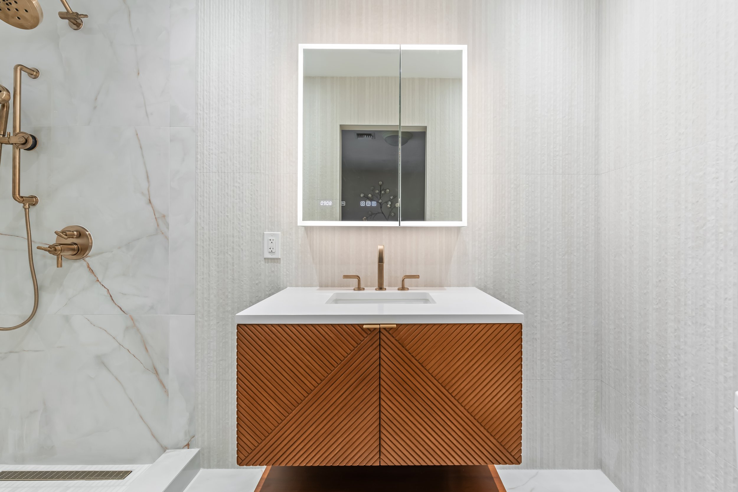 Modern bathroom with marble shower wall with gold fixtures, a sleek mirror cabinet with LED lighting, a white rectangular sink, a wood cabinet under the sink, and light-colored textured wallpaper.