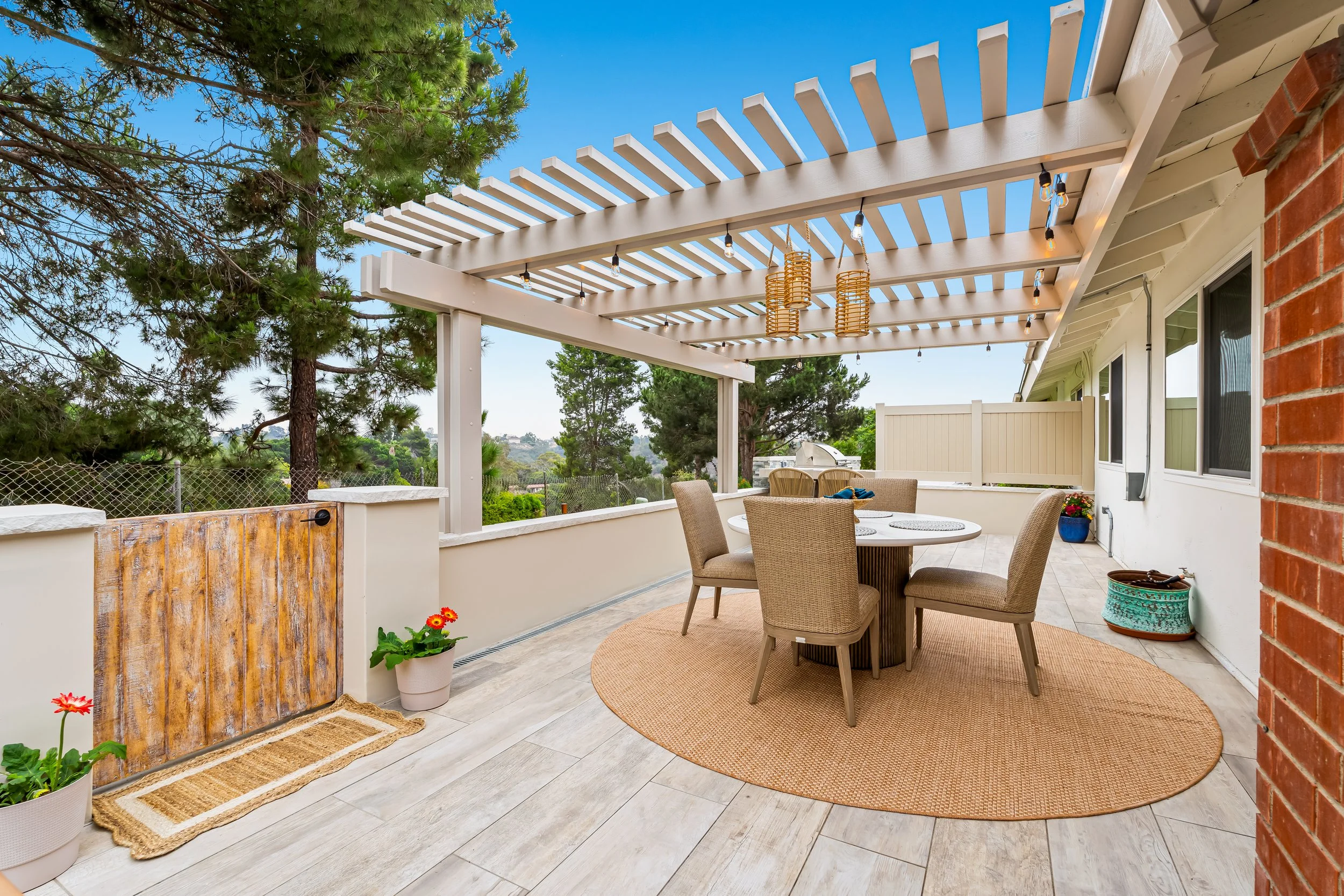 A covered outdoor patio area with patio furniture, potted plants, a string of lights, and a view of trees and sky.
