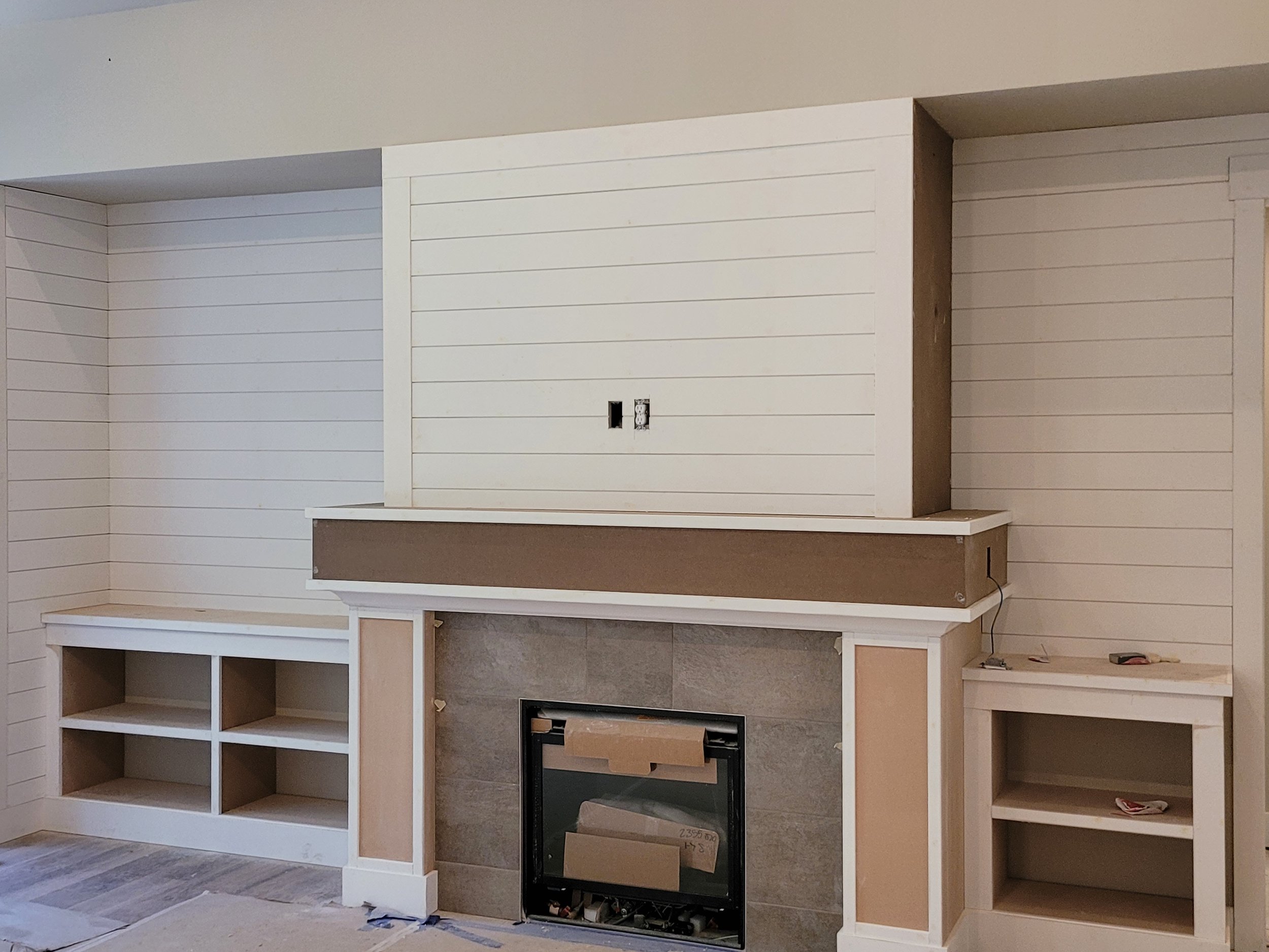 Room under construction with built-in fireplace and shelving unit, partially painted walls, and construction materials on the floor.