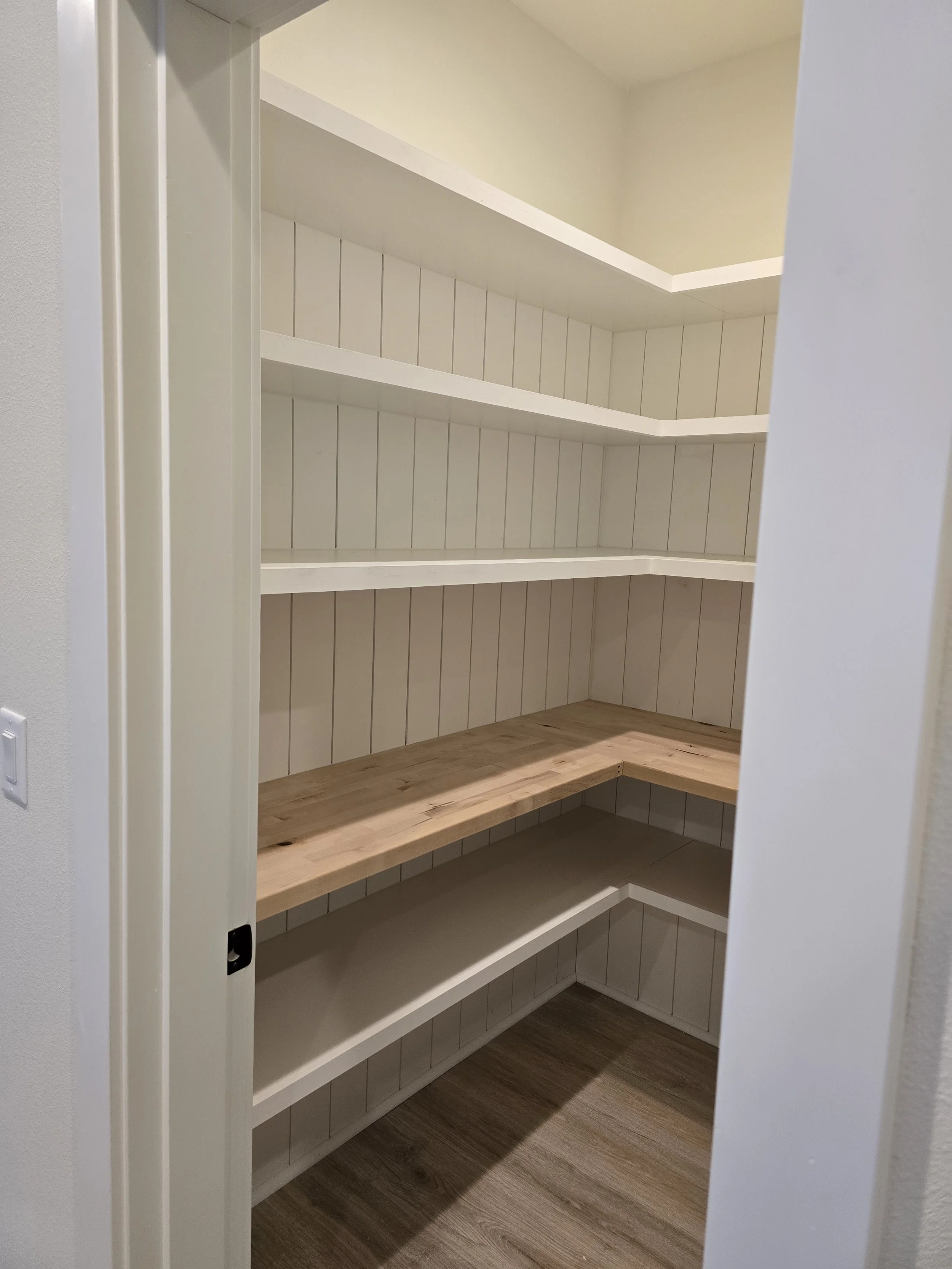 Empty white pantry with wooden shelves and wood flooring, viewed through partially open door.